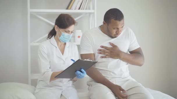 Mujer con máscara facial escribiendo en documentos como hombre tosiendo. Médico femenino caucásico serio seguro que llena los síntomas del paciente varón enfermo en la historia clínica. Visita del médico a la pandemia de Covid-19. — Vídeo de stock