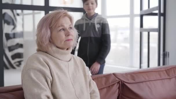 Beautiful mature woman sitting on couch at home as cute Caucasian boy and girl coming and hugging grandmother. Portrait of happy grandchildren visiting grandparent on holidays. — Stock Video