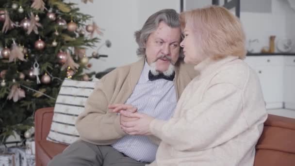Milující starý bělošský pár držící se za ruce a mluvící o Vánocích. Portrét bezstarostných důchodců, kteří si na Silvestra povídají doma. Romantický starší muž a žena sedí na gauči a mluví.