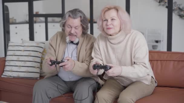 Alegre absorbido senior hombre y mujer jugando videojuegos con consolas de juegos. Retrato de una alegre pareja de ancianos divirtiéndose en casa en la víspera de Año Nuevo. Relajados marido y mujer disfrutando de vacaciones de Navidad. — Vídeo de stock