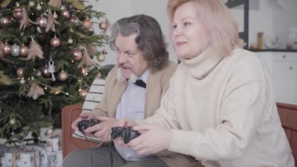 Vista de ángulo lateral de alegre pareja de mayores caucásicos jugando videojuegos en la víspera de Año Nuevo en casa. Relajados jubilados felices divirtiéndose como jugando en Navidad. Ocio y estilo de vida. — Vídeos de Stock