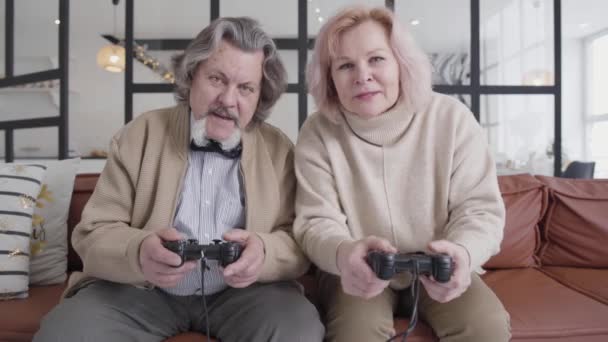 En medio de una divertida pareja de ancianos jugando videojuegos. Retrato de los alegres esposos caucásicos usando consolas de juegos y riendo. Jubilados positivos divirtiéndose en Navidad en casa. — Vídeo de stock