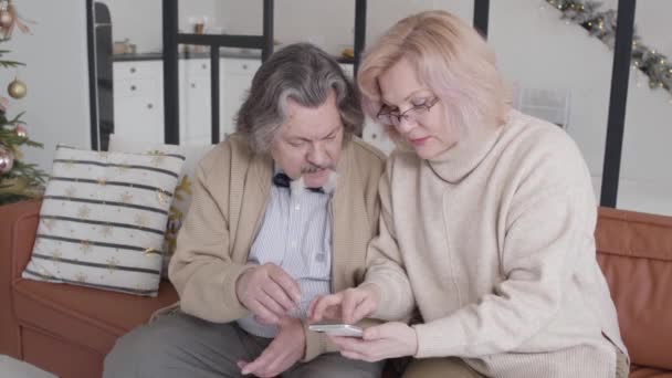 Seguro de alto nivel hermosa mujer tocando el teléfono inteligente y hablando con el hombre. Retrato de la encantadora esposa caucásica explicando las tecnologías modernas al marido en la víspera de Navidad. Cuidado y apoyo. — Vídeos de Stock