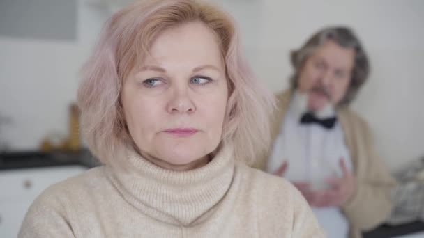 Close-up of beautiful senior woman with blurred angry man shouting at the background. Portrait of frustrated Caucasian wife tired of husbands nagging. Conflict of old married couple. — Stock Video