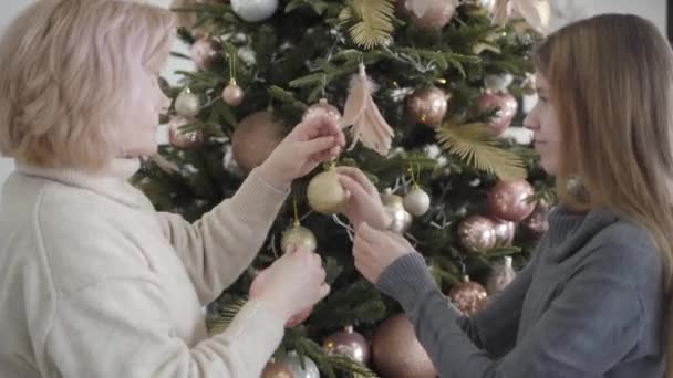 Visszapillantás aranyos tizenéves lány segít idősebb nő dekoráció az újévi fa. Mosolygó unoka ünnepli a karácsonyt a kaukázusi nagymamával. Egység és ünneplés koncepció. — Stock videók