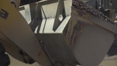 Büyük endüstriyel traktör dolu kovayla kameranın önünden geçiyor. Üretim alanında ezilmiş taşla dolu bir yük var. Fabrika makineleri gün ışığında çalışıyor..