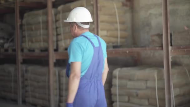 Κάμερα ακολουθεί βέβαιος Καυκάσιος άνδρας περπατώντας στις εγκαταστάσεις αποθήκευσης του χώρου παραγωγής. Ένας ενήλικας εργάτης με στολή και κράνος στο χώρο εργασίας στο εργοστάσιο. Εργοστάτης της βιομηχανίας παραγωγής σκυροδέματος — Αρχείο Βίντεο