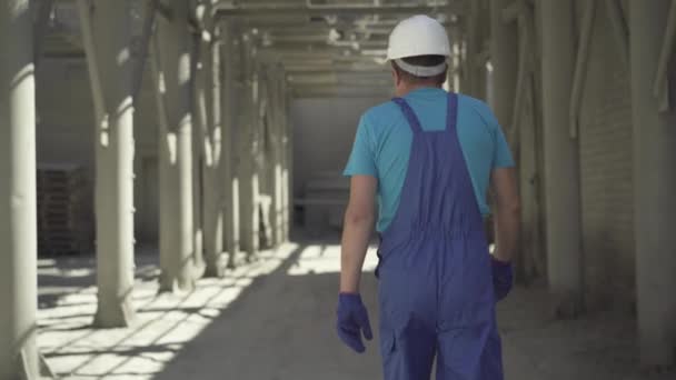 Pohled zezadu na dospělého bělocha v uniformě a helmě kráčejícího po staveništi za slunečního svitu. Kamera sleduje mužského pracovníka procházky na pracovišti za slunečného letního dne. — Stock video