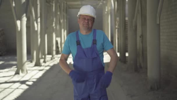 Уверенный позитивный человек в форме и шлеме скрещивает руки и улыбается в камеру. Портрет кавказского строителя, позирующего на стройке в солнечных лучах в солнечный летний день. — стоковое видео