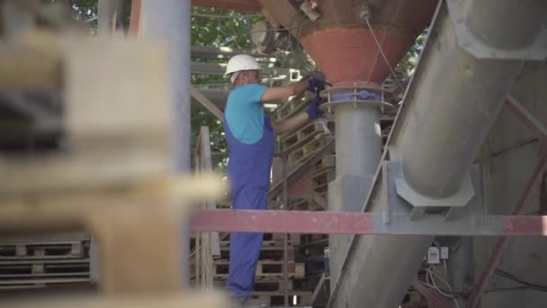 Vue d'ensemble de l'équipement de réglage des ouvriers d'usine à l'extérieur de l'usine. Homme caucasien professionnel en uniforme et casque travaillant avec un convoyeur à vis en ciment et laissant. — Video