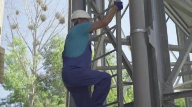 Fabrika işçisinin devasa paslanmaz çelik beton depolama silosunun üzerinde merdivenleri tırmanırken yan görüntüsü. Kamera, beyaz üniformalı ve kasklı adamı beton üretim tesisinde çalışırken izliyor..