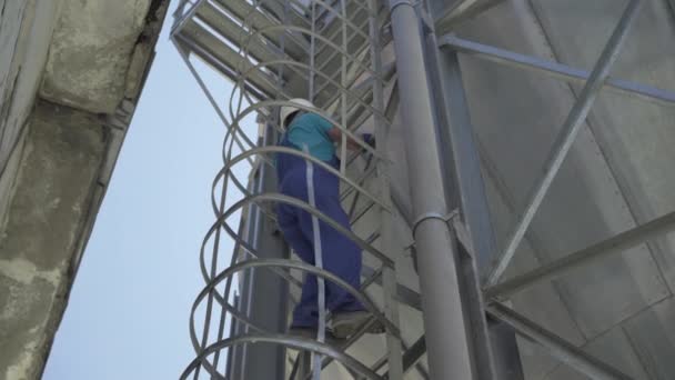 Vista inferior de un trabajador caucásico que sube por la escalera en una enorme torre de metal. Hombre profesional de confianza en uniforme y casco de seguridad trabajando en silo de almacenamiento de cemento al aire libre. — Vídeos de Stock