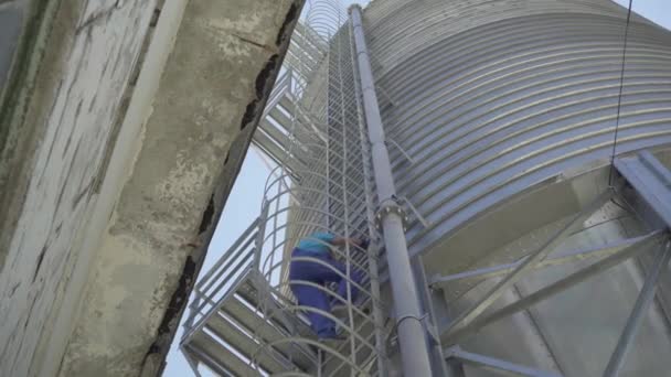 Gros plan de l'homme en uniforme et casque de sécurité marchant sur un énorme silo de stockage de ciment. Immense tour en acier inoxydable avec ouvrier d'usine caucasien grimpant en utilisant l'échelle. — Video