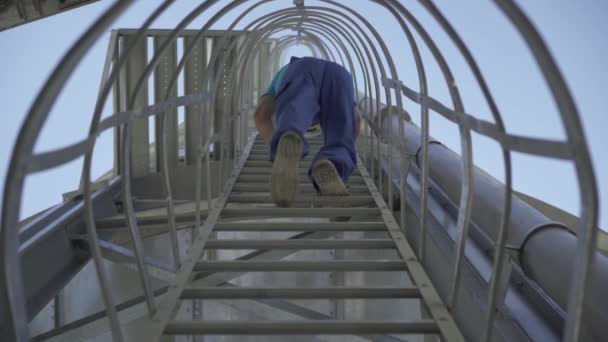 Vista inferior do trabalhador da fábrica em uniforme azul descendo a escada na enorme torre de armazenamento. Homem caucasiano irreconhecível no silo de cimento ao ar livre com céu azul claro no fundo. — Vídeo de Stock