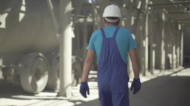 Blanke man in uniform en helm lopen op beton productielocatie met cement mixer truck rijden. Mannelijke blanke werknemer controleert productie op fabriek of fabriek. — Stockvideo