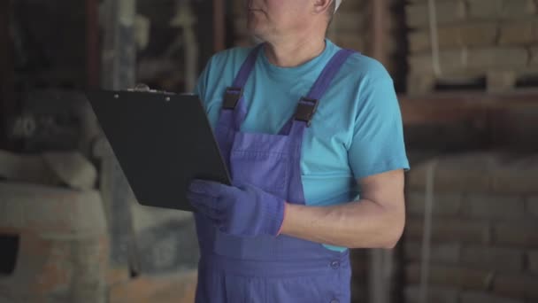 倉庫に立つ文書で書かれた青い制服と作業手袋で認識できない労働者。工場や工場の倉庫で働く男性白人従業員. — ストック動画