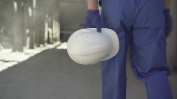 Närbild av vit hjälm i händerna på en vit man som går på byggplatsen i solljus. Manlig byggare i blå uniform och arbetshandskar på byggarbetsplatsen utomhus. — Stockvideo