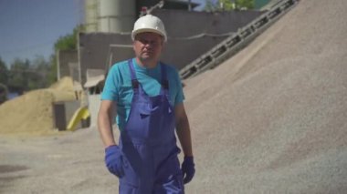 Mavi üniformalı, kasklı, kendinden emin beyaz bir adamın portresi. Toptan malzeme yığınları boyunca üretim sahasında yürüyor. Güneşli bir günde iş yerinde çalışan profesyonel bir fabrika işçisi..