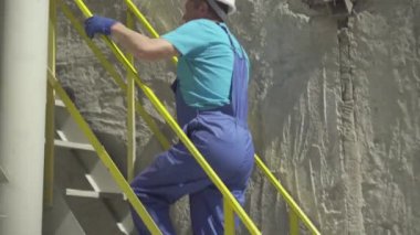 Üniformalı beyaz adamın üretim alanında merdivenleri çıkarken geniş açılı görüntüsü. Kasklı profesyonel erkek işçi ve çimento üretim fabrikasında çalışan eldivenler..
