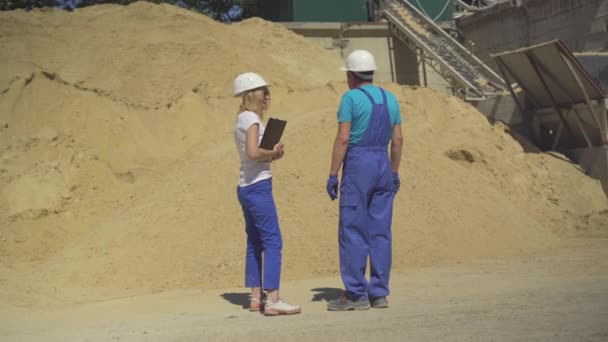 Brett skott av professionella kvinnliga inspektörer och manliga arbetare som står på tillverkningsplatsen framför enorma sandhög och pratar. Vit man och kvinna i skyddshjälmar på cementfabrik. — Stockvideo