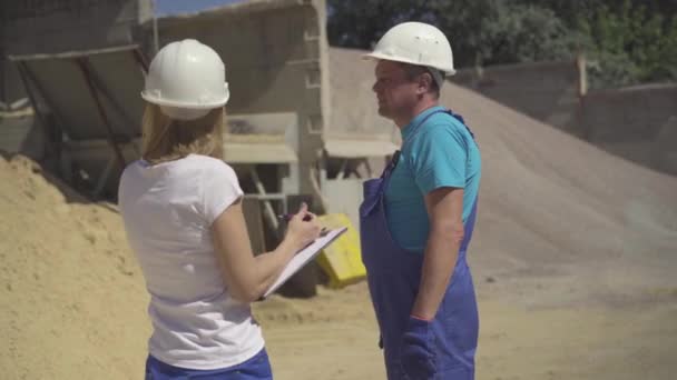 Un colpo medio di donna caucasica sicura di se 'con documenti che parlano con un uomo adulto all'aperto sul luogo di produzione. Operaio e ispettore donna che discutono di produzione di calcestruzzo. — Video Stock