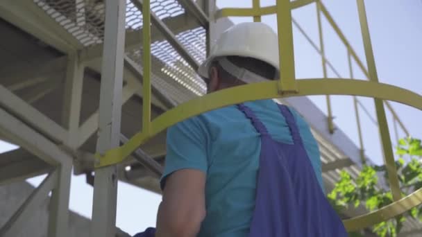 La cámara sigue los pasos del trabajador de la fábrica que sube la escalera al aire libre en el día soleado. Hombre caucásico profesional adulto en uniforme azul y casco de seguridad blanco en el sitio de fabricación. — Vídeo de stock