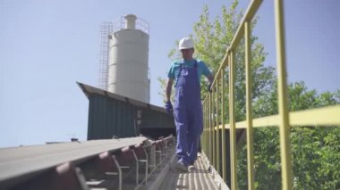 Profesyonel fabrika işçisinin geniş açılı portresi açık havada konveyör yükleyip vanaları kontrol ediyor. Beton üretim tesisinin otomatik çimento yükleme ekipmanlarının elle teftişi.