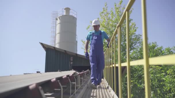 Wide shot portrait of professional factory worker walking along loading conveyor outdoors and checking valves. Manual inspection of automated cement loading equipment on concrete manufacturing plant. — Stock Video