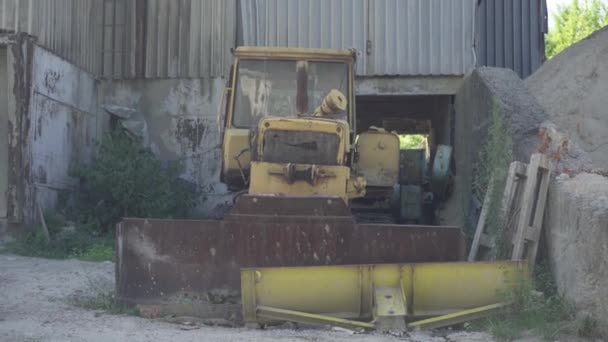 Široký pohled zepředu na rezavý rozbitý traktor s vědrem stojícím na troskách továrny. Staré těžké průmyslové stroje na opuštěné továrně venku. — Stock video