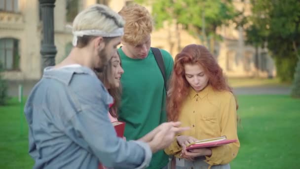 Grupo de colegas de grupo positivos explicando lição de casa para encantadora ruiva mulher caucasiana. Quatro homens e mulheres alegres em pé ao ar livre no pátio da universidade ensolarado e falando. — Vídeo de Stock