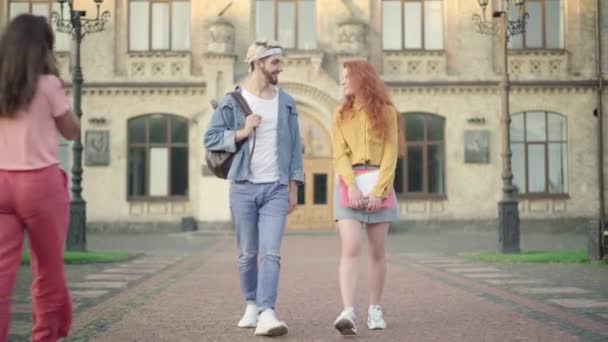 Beau jeune homme flirtant avec belle rousse femme comme marcher après les cours dans la cour de l'université. Portrait d'étudiants souriants et attrayants se promenant comme des amis saluant en arrière-plan. — Video