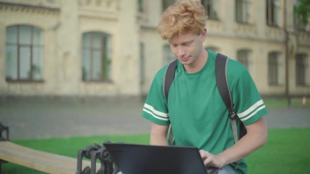 Encantadora pelirroja uniéndose a un hombre caucásico concentrado sentado en el banco del campus y usando una computadora portátil. Retrato de dos estudiantes universitarios confiados que estudian en la universidad. Concepto educativo. — Vídeo de stock