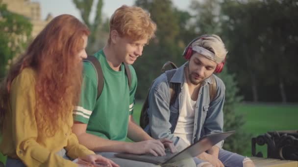 Troje zrelaksowanych studentów siedzących na kampusie i surfujących po Internecie. Rudowłosy biały mężczyzna wpisując na klawiaturze laptopa jako blond facet słuchając muzyki w słuchawkach i kobieta mówi. — Wideo stockowe