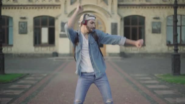 Портрет веселого хлопця в навушниках, який ходить і танцює, коли студенти проходять на задньому плані. Насолоджуйся відпочинком після занять у університетському містечку.. — стокове відео