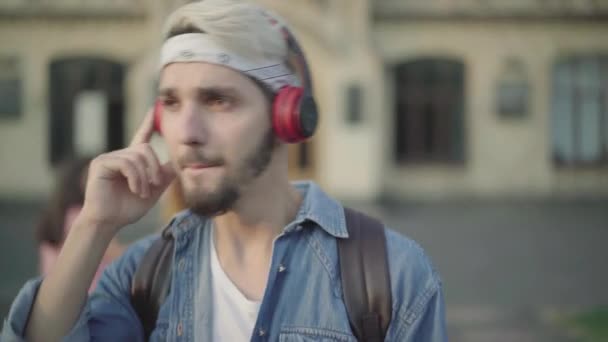 Retrato de close-up de belo jovem barbudo em fones de ouvido andando ao ar livre e cantando. Estudante desfocado passando em segundo plano. Estudante caucasiano masculino confiante no quintal da faculdade ou da universidade. — Vídeo de Stock