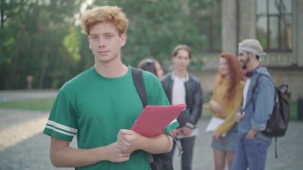 Portrait de jeune homme rousse caucasien confiant posant sur le campus de l'université avec des camarades flous parlant en arrière-plan. Beau gars intelligent regardant la caméra et souriant. — Video