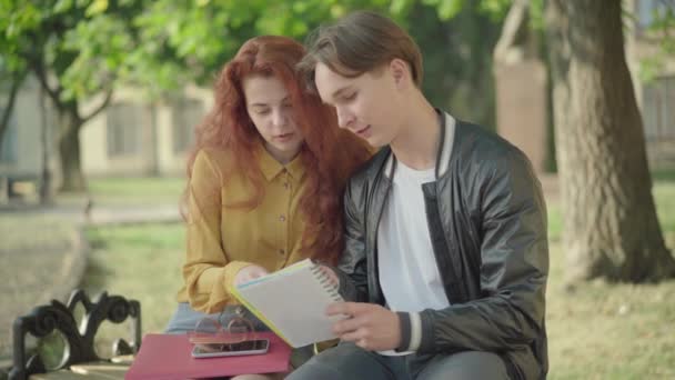 Portrait médian du jeune homme et de la jeune femme caucasiens assis sur le campus et parlant. Heureux étudiants discutant des devoirs à l'extérieur. Éducation et amitié. — Video