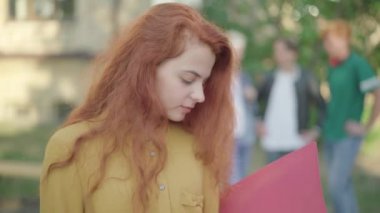 Genç, çekici, kızıl saçlı kadının kameraya dönüp gülümsemesinin yakın çekimi. Arka planda bulanık insanlarla kampüste poz veren güzel beyaz üniversite öğrencisinin portresi..