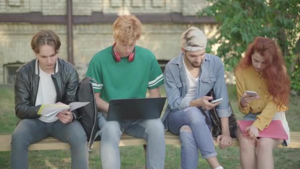 Fire koncentrerede universitetsstuderende surfer på internettet og laver lektier på campus. Portræt af selvsikker fokuseret kaukasiske mænd og kvinder sidder på bænken udendørs. Livsstil koncept. – Stock-video
