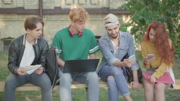 Hombre seguro positivo mostrando la pantalla del teléfono inteligente a los compañeros de grupo al aire libre. Cuatro estudiantes universitarios caucásicos navegando por Internet después de estudiar en el patio de la universidad. Estilo de vida moderno. — Vídeo de stock