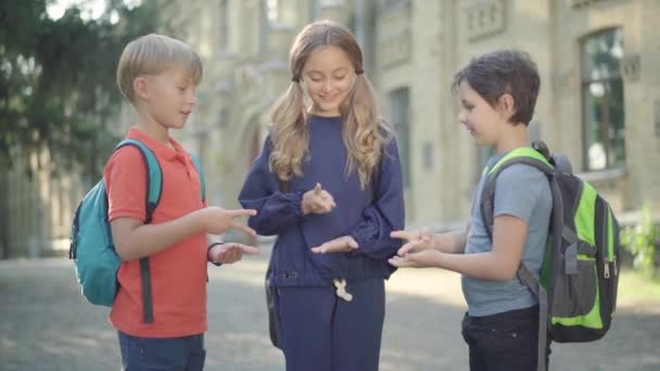 Trois enfants joyeux détendus debout sur la cour de l'école et jouant pierre-papier-ciseaux. Portrait de joyeux écoliers caucasiens insouciants et écolière s'amuser en plein air le jour ensoleillé. — Video