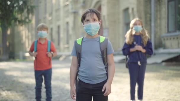 Portrait de petit garçon brune masqué debout au soleil dans la cour de l'école avec des camarades de classe flous à l'arrière-plan. Concept de distanciation sociale dans le processus d'éducation Covid-19. — Video