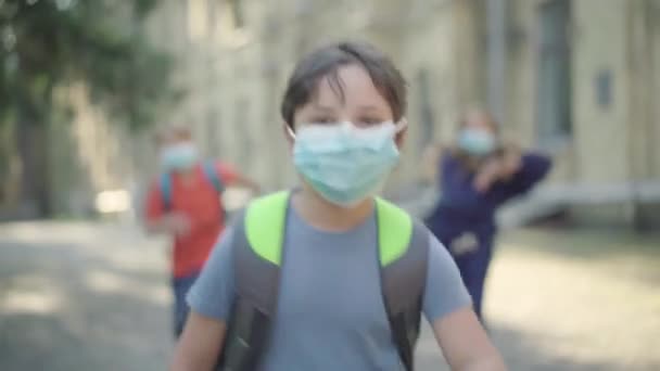 Porträtt av bekymmerslös skolpojke i ansiktsmask löper längs skolgården i solljus med suddiga klasskamrater i bakgrunden. Glada kaukasiska barn har roligt efter lektionerna på Covid-19 pandemi. — Stockvideo