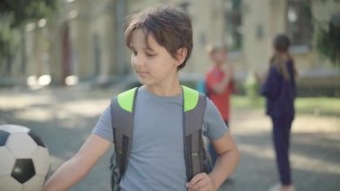 Güneşli okul bahçesinde futbol ya da futbol topuyla poz veren kendinden emin ortaokul öğrencisinin portresi arka planda el çırpan bulanık sınıf arkadaşları gibi. Beyaz çocuk kameraya gülümsüyor..