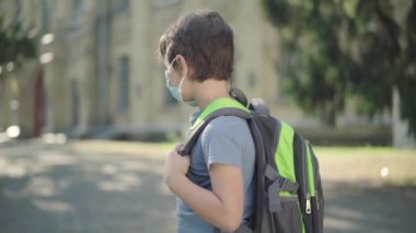 Sırt çantalı, maske takan üzgün okul çocuğu kameraya dönüyor. Güneşli okul bahçesinde dikilen üzgün beyaz çocuğun portresi. Covid-19 salgını sırasında ortaokulda okuyan tatlı esmer çocuk..