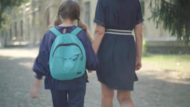 Kamera följer brunett liten flicka promenader längs solig skolgård med oigenkännlig mor. Baksidan av skolflicka promenader med förälder efter lektionerna. Utbildning och enighet. — Stockvideo