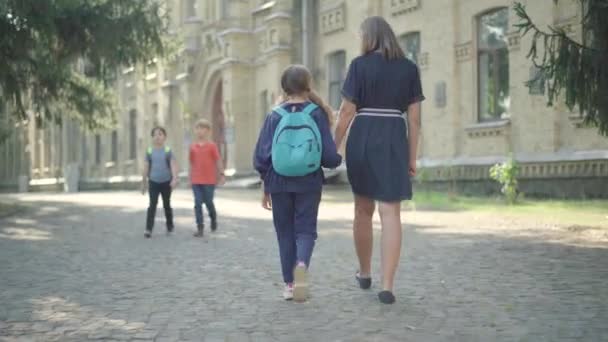 Bred bild av mor och dotter som går längs solig skolgård och pratar. Bakåt syn på positiv skolflicka med ryggsäck promenader med förälder utomhus. Grundskolans utbildningskoncept. — Stockvideo