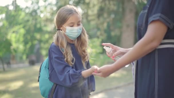 学校の庭で女子高生の手に消毒剤を噴霧する認識できない女性。顔のマスクのかわいい女の子は手のひらを消毒し、逃げる。Covid-19パンデミック時の教育. — ストック動画