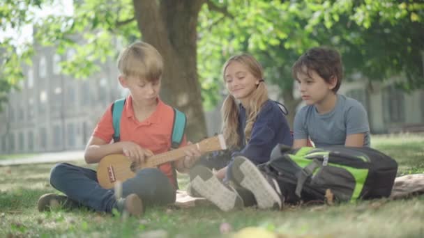 Crianças em idade escolar despreocupada sentadas no prado ensolarado no pátio da escola à luz do sol. Loira caucasiano menino jogando ukulele e amigos batendo palmas. Passatempo e estilo de vida dos alunos do ensino médio. — Vídeo de Stock