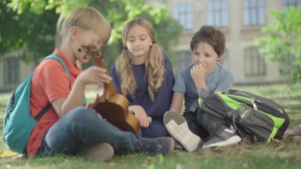 Niños caucásicos relajados sentados en el patio de la escuela soleado y jugando ukelele. Retrato de estudiantes y amigos de secundaria despreocupados descansando después de estudiar al aire libre. Concepto de amistad. — Vídeos de Stock